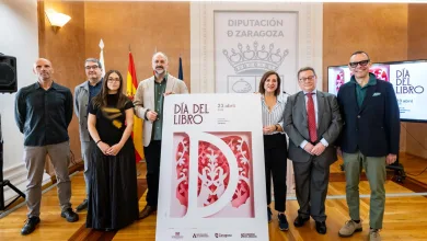 Eight people pose behind a tall Día del Libro poster at a Zaragoza event, with flags and a crest in the background in a wood-paneled room.