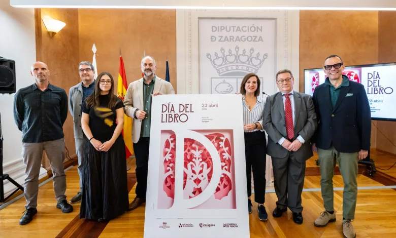 Eight people pose behind a tall Día del Libro poster at a Zaragoza event, with flags and a crest in the background in a wood-paneled room.