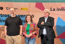 Three people posing at a film festival backdrop: a man in a black t‑shirt on the left, a woman in a green shirt with a colorful floral jacket in the center, and a man in a dark suit with arms crossed on the right.