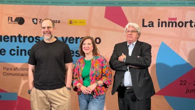 Three people posing at a film festival backdrop: a man in a black t‑shirt on the left, a woman in a green shirt with a colorful floral jacket in the center, and a man in a dark suit with arms crossed on the right.