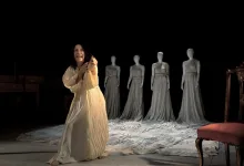 Theatrical scene: a woman in a cream flowing gown center stage, gesturing forward, with five gray-dressed mannequins lined up behind her on a draped floor.