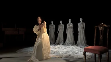 Theatrical scene: a woman in a cream flowing gown center stage, gesturing forward, with five gray-dressed mannequins lined up behind her on a draped floor.