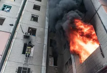 Firefighters on a ladder rescue a person from a building window as a large fire and dark smoke erupt from a tower next door.