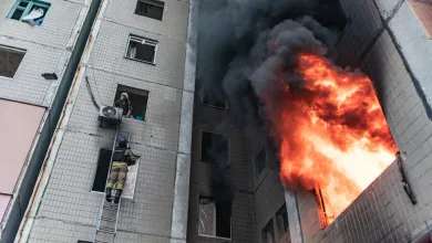 Firefighters on a ladder rescue a person from a building window as a large fire and dark smoke erupt from a tower next door.