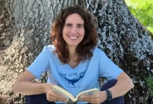 Woman sitting against a large tree trunk outdoors, smiling while reading an open book.