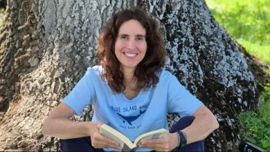 Woman sitting against a large tree trunk outdoors, smiling while reading an open book.