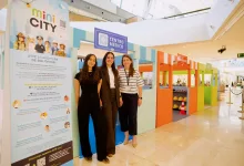 Three smiling women pose at a colorful kids’ exhibit with a large mini CITY poster and teal-orange structures nearby in a shopping mall.