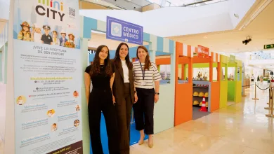 Three smiling women pose at a colorful kids’ exhibit with a large mini CITY poster and teal-orange structures nearby in a shopping mall.