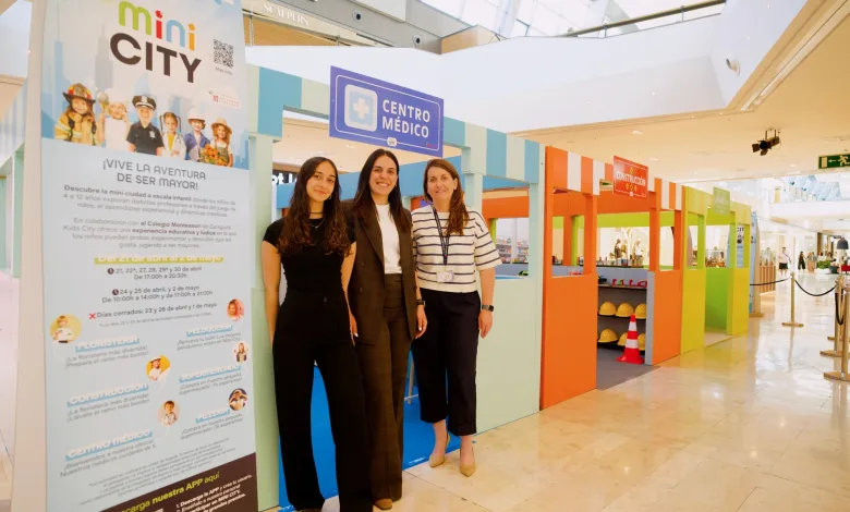 Three smiling women pose at a colorful kids’ exhibit with a large mini CITY poster and teal-orange structures nearby in a shopping mall.