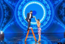 Man in a suit helps a woman in a floral dress up on an orange ladder on a blue, ornate stage backdrop.