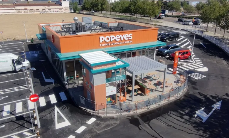 Aerial view of a Popeyes Louisiana Chicken restaurant with an orange sign, teal awnings, and an outdoor seating patio surrounded by a circular drive-through lane.