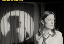 Black-and-white photo of a woman wearing a striped tie, facing right on a stage with dark curtains behind her.
