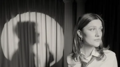 Black-and-white photo of a woman wearing a striped tie, facing right on a stage with dark curtains behind her.