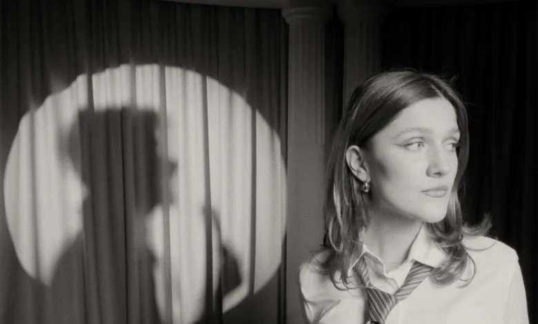 Black-and-white photo of a woman wearing a striped tie, facing right on a stage with dark curtains behind her.