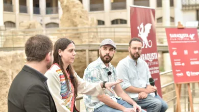 Panelists sitting in a row outdoors during a discussion, with red festival banners in the background and microphones visible on their shirts.