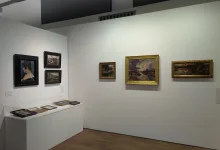 Gallery interior with framed paintings on white walls and a glass display case containing small framed works on the left.