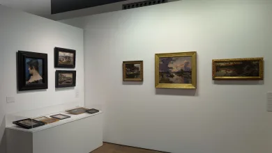 Gallery interior with framed paintings on white walls and a glass display case containing small framed works on the left.