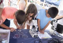 Group of kids at a science fair leaning over a table with microscopes, examining slides and taking notes.