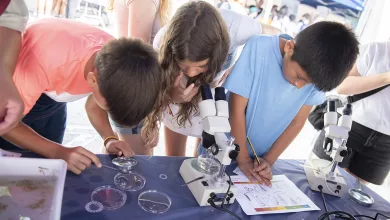 Group of kids at a science fair leaning over a table with microscopes, examining slides and taking notes.