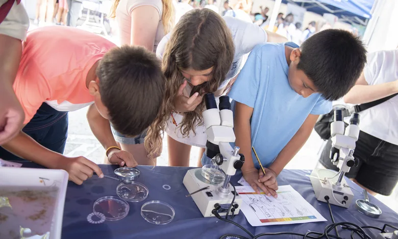 Group of kids at a science fair leaning over a table with microscopes, examining slides and taking notes.