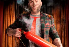 Man on a stage wearing a plaid blazer and red tie holds a large red cylindrical prop emitting smoke and small flames, with theater curtains in the background.