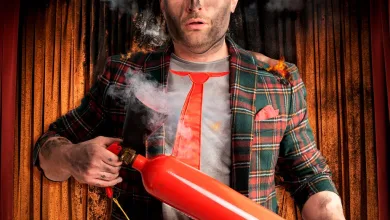 Man on a stage wearing a plaid blazer and red tie holds a large red cylindrical prop emitting smoke and small flames, with theater curtains in the background.