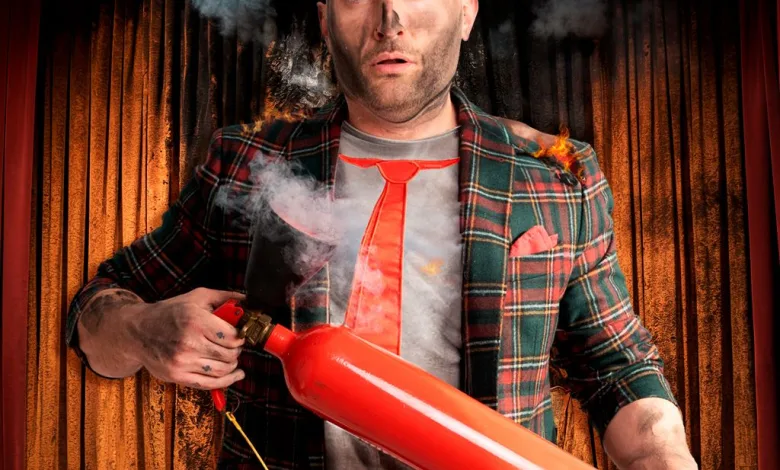 Man on a stage wearing a plaid blazer and red tie holds a large red cylindrical prop emitting smoke and small flames, with theater curtains in the background.