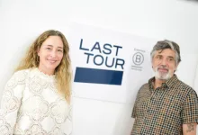 Two smiling people posing in front of a 'LAST TOUR' sign; woman on left wearing white lace top, man on right in patterned shirt.