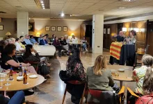 Audience seated around round tables listening to two speakers at a small elevated lectern with a striped cloth and drinks on the tables used for a talk event in a lounge area of a venue.