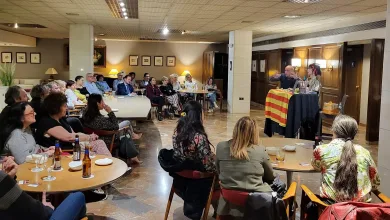 Audience seated around round tables listening to two speakers at a small elevated lectern with a striped cloth and drinks on the tables used for a talk event in a lounge area of a venue.