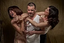 Man in a white sleeveless shirt is restrained from behind by two women in a staged, dramatic pose.