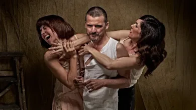 Man in a white sleeveless shirt is restrained from behind by two women in a staged, dramatic pose.