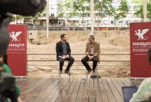 Two men in jackets sit on stools on a wooden stage at a film festival, with red banners showing a dragon logo in the background.