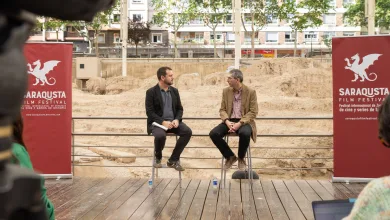 Two men in jackets sit on stools on a wooden stage at a film festival, with red banners showing a dragon logo in the background.