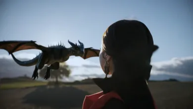 Person in a dark cloak watches a small blue dragon soaring over a rural landscape.