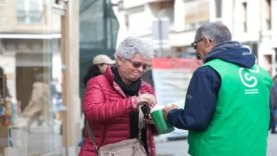 An elderly woman in a red jacket accepts a donation container from a man wearing a green volunteer vest on a city street.
