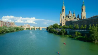 Wide river with a stone arch bridge and a cityscape featuring ornate cathedral towers along the right bank under a bright blue sky.