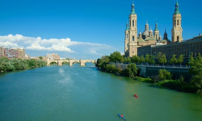 Wide river with a stone arch bridge and a cityscape featuring ornate cathedral towers along the right bank under a bright blue sky.