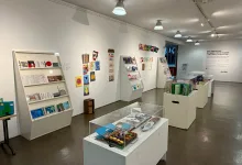 Interior view of a bright art gallery with white walls, angled display shelves filled with colorful small artworks and books.