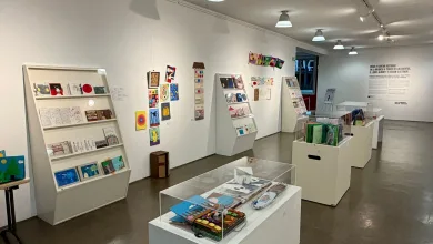 Interior view of a bright art gallery with white walls, angled display shelves filled with colorful small artworks and books.