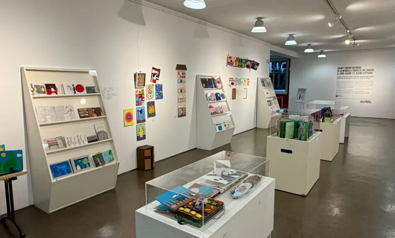Interior view of a bright art gallery with white walls, angled display shelves filled with colorful small artworks and books.