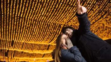 Two friends pose for a selfie beneath warm string lights at night, smiling as one points upward.