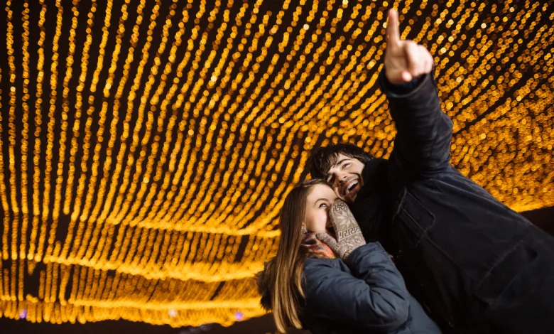Two friends pose for a selfie beneath warm string lights at night, smiling as one points upward.