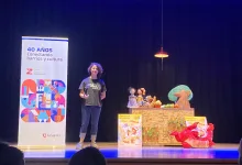 Presenter with Curly Hair on a Stage Beside a Table of Puppets and a Tall Banner Reading 40 AÑos Conectando Barrios Y Cultura Zaragoza   Qué Hacer En Zaragoza Zaragozala   Qué Hacer En Zaragoza