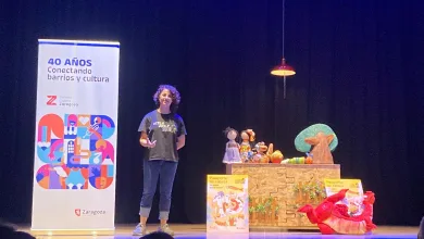 Presenter with Curly Hair on a Stage Beside a Table of Puppets and a Tall Banner Reading 40 AÑos Conectando Barrios Y Cultura Pasaporte Sin Cabeza Lleva El Teatro Familiar a Más De 1300 Personas Zaragozala   Qué Hacer En Zaragoza