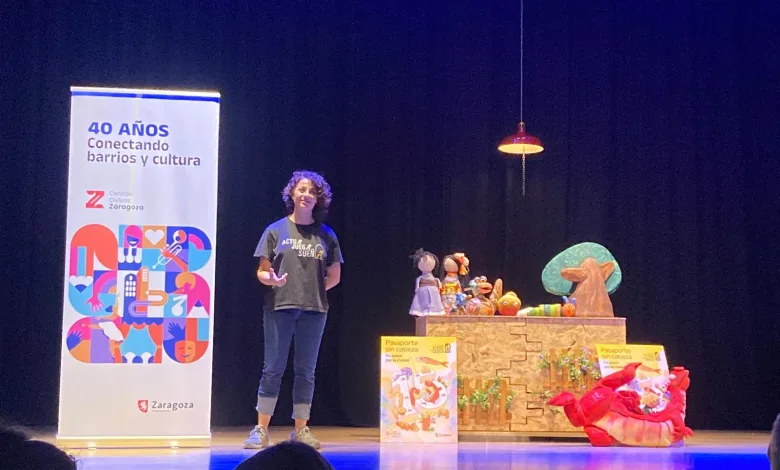 Presenter with Curly Hair on a Stage Beside a Table of Puppets and a Tall Banner Reading 40 AÑos Conectando Barrios Y Cultura Pasaporte Sin Cabeza Lleva El Teatro Familiar a Más De 1300 Personas Zaragozala   Qué Hacer En Zaragoza