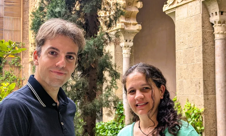 Two people pose outdoors in front of an ornate carved stone archway with greenery nearby.