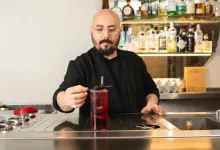Bartender with glasses and beard in a black shirt garnishes a tall red cocktail with a blackberry skewer at a bar.