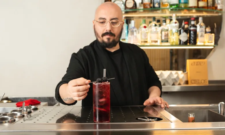 Bartender with glasses and beard in a black shirt garnishes a tall red cocktail with a blackberry skewer at a bar.