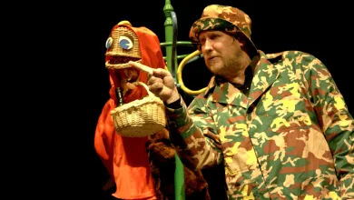 Man in Camouflage Jacket and Hat Points Toward a Wicker Basket Puppet Wearing a Red Hood on a Dark Stage Background Zaragoza   Qué Hacer En Zaragoza Zaragozala   Qué Hacer En Zaragoza